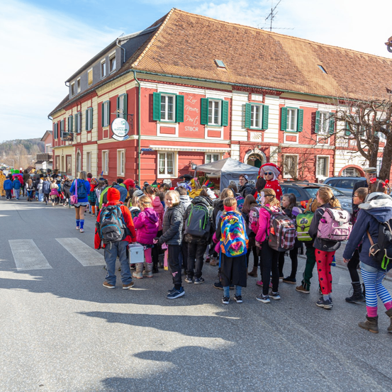 Eine Gruppe von Kindern mit Rucksäcken steht auf der Straße vor einem großen Gebäude mit roten Wänden und grünen Fensterläden. Eine Person in Santa-Claus-Kleidung ist anwesend.