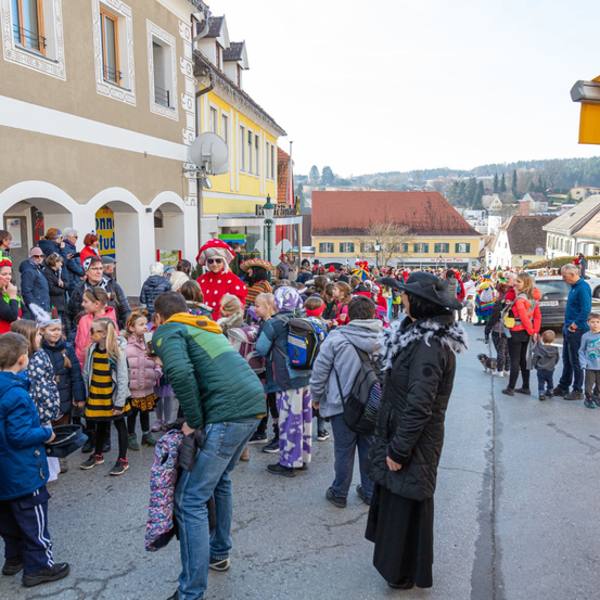 Eine Menschenmenge, darunter auch Kinder, versammelt sich auf einer Straße. Einige sind in Kostümen gekleidet. Gebäude säumen die Straße, und in der Ferne sind Autos und Bäume zu sehen.
