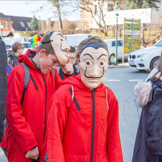 Zwei Jungen in roten Overalls und Masken gehen auf der Straße, einer berührt seine Maske. Hinter ihnen steht eine Person in der Nähe eines geparkten weißen Autos. Im Hintergrund befinden sich Gebäude, Bäume und ein Zaun.