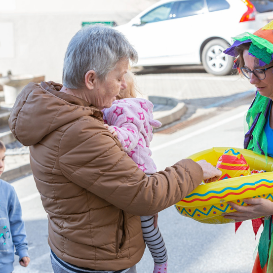 Eine ältere Frau, die ein junges Kind trägt, reicht einer Person in bunter Kleidung eine aufblasbare Schwimmhilfe. Im Hintergrund ist ein weißer Wagen geparkt.