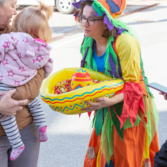 Eine Frau in einem bunten Kostüm hält eine große Süßigkeiten-Verpackung und interagiert mit einem Kleinkind in einem rosa Mantel.