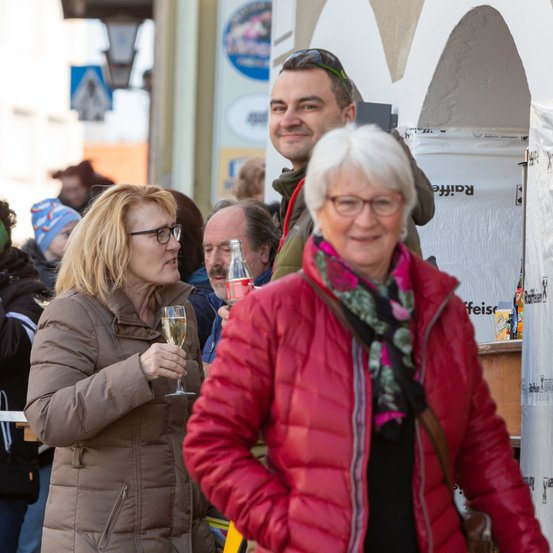 Zwei Frauen, eine mit einem Weinglas, stehen neben einem Mann mit Brille und Schal. Der Mann hat eine Sonnenbrille auf dem Kopf. Hinter ihnen stehen weitere Menschen und unterhalten sich. Ein Gebäude ist im Hintergrund.