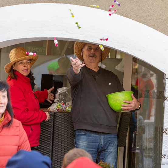 Ein Mann und eine Frau stehen unter einem Bogen, der Mann hält eine grüne Schüssel voller Süßigkeiten. Konfetti fällt von oben. Eine weitere Frau in einer roten Jacke schaut zu.