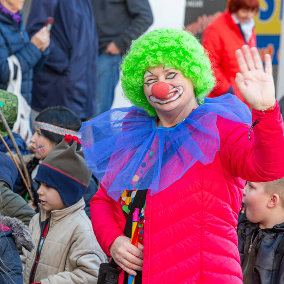 Ein Clown in einem roten Kostüm winkt einer Menge zu, wobei Kinder zusehen, einer hält einen Stock und trägt eine farbenfrohe Kopfbedeckung.