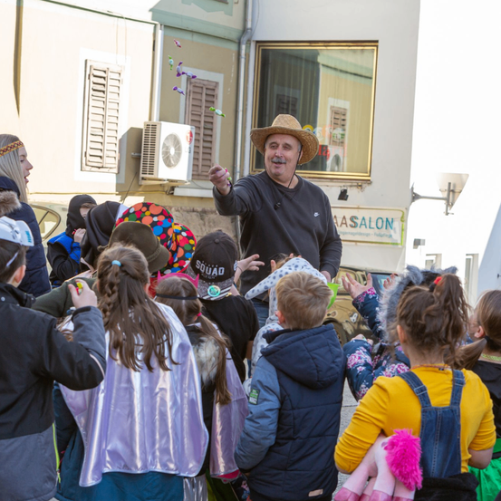 Ein Mann mit einem Strohhut steht vor einer Gruppe von Kindern, wahrscheinlich als Unterhaltung. Die Kinder tragen verschiedene Kostüme und einige halten Requisiten. Hinter dem Mann befindet sich ein Gebäude mit einer Klimaanlage und einem Schild mit der Aufschrift 'As Salon'.