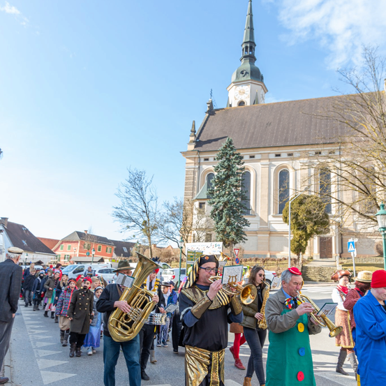Eine Blaskapelle mit Blechinstrumenten spielt auf einer Straße vor einer Kirche. Einige Menschen sind in Kostümen, und es gibt Bäume und Straßenlaternen in der Umgebung.