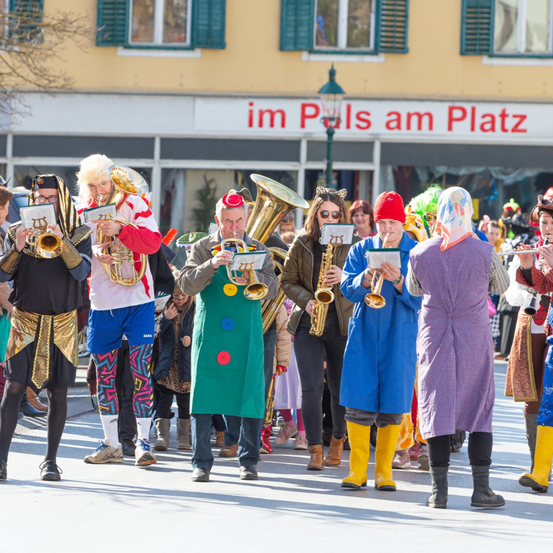 Eine Gruppe von Musikern in bunten Kostümen spielt Blech- und Holzblasinstrumente bei einem Umzug vor einem Gebäude mit dem Text 'im Pols am Platz'.