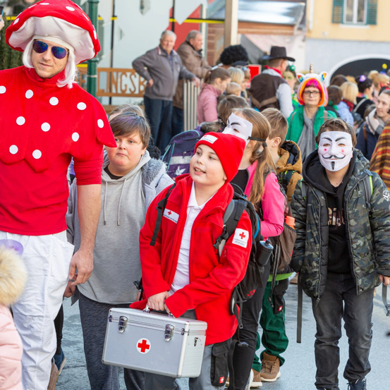 Eine Gruppe von Menschen in Kostümen zieht bei einem Umzug. Ein Mann trägt ein rotes Punktepilzkostüm, während ein Junge eine rote Kreuz-Tasche hält.
