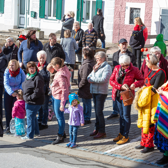 Eine Menschenmenge, darunter ein kleines Kind, versammelt sich auf einer Straße. Einige tragen Mützen, Brillen und Stiefel. Hinter ihnen sind mehrere Gebäude mit Fenstern und Türen zu sehen.