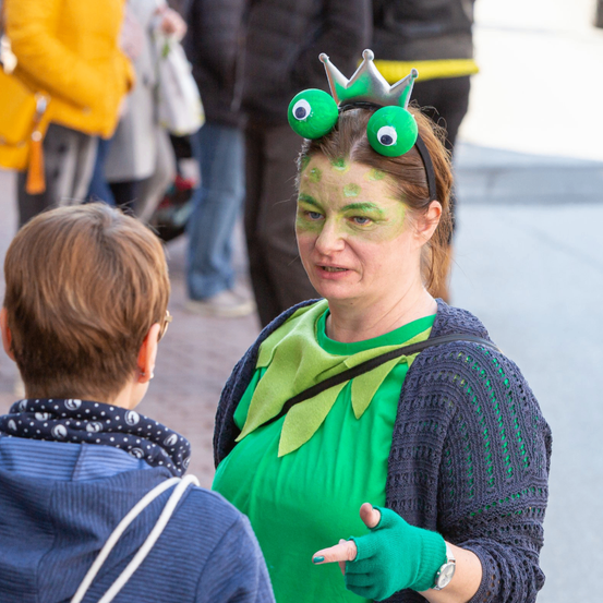 Eine Frau in einem Froschkostüm spricht mit einem jungen Kind in einem städtischen Umfeld. Die Frau hat grünes Gesichtsmake-up, eine Krone und grüne Handschuhe.