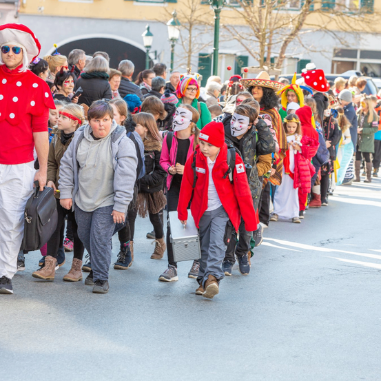 Eine Menschenmenge, bestehend aus Kindern und Erwachsenen, ist in verschiedenen Kostümen gekleidet und läuft auf einer Straße. Einige tragen Masken, und ein Kind hält eine Aktentasche. Im Hintergrund sind Gebäude und Bäume zu sehen.