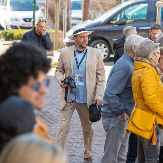Ein Mann mit einer Kamera steht in einer Menschenmenge, trägt einen Hut und eine Lanyard. In der Nähe versammeln sich mehrere Personen, einige tragen Brillen und Jacken. Ein Van und ein Auto sind im Hintergrund geparkt.