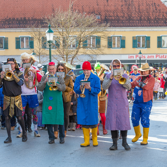 Eine Gruppe von Musikern in bunten Kostümen steht in der Straße und spielt Trompeten und Tuben. Einige tragen Stiefel und Hüte.