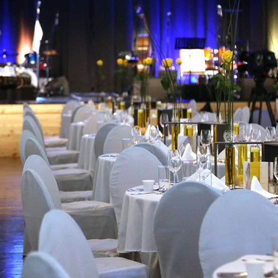 Ein großer Speisesaal mit weißen Stühlen und Tischen, jeder mit Weingläsern, Tellern und gelben Blumen besetzt. Eine Bühne mit Trommeln, einer Fahne und Musikinstrumenten im Hintergrund.