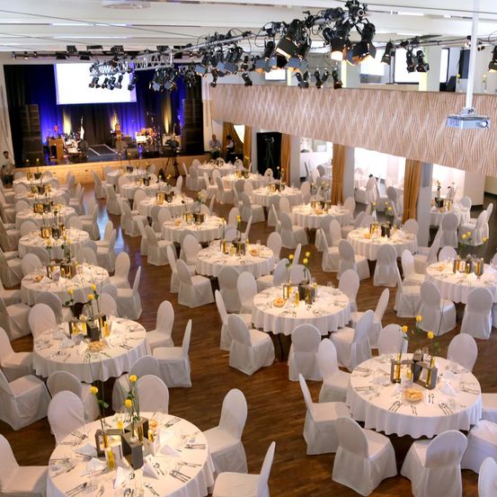 Ein großer Bankettsaal ist für eine Veranstaltung vorbereitet mit weißen Tischen und Stühlen. Jeder Tisch hat einen gelben Blumen-Zentrenplatz. Eine Bühne mit Leinwand und Lautsprechern ist im Hintergrund eingerichtet.