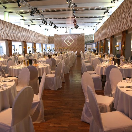Ein Bankettsaal ist mit weißen Stühlen und Tischen eingerichtet. Jeder Tisch hat eine weiße Tischdecke und gelbe Blumen. Die Wände sind mit einem goldenen Design verziert. Scheinwerfer sind an der Decke installiert.