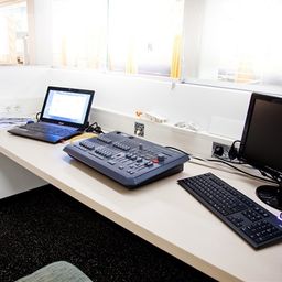 Ein Büroschreibtisch ist mit einem Laptop, einem Monitor, einer Tastatur, einer Maus und einem Lichtsteuerungspanel ausgestattet.