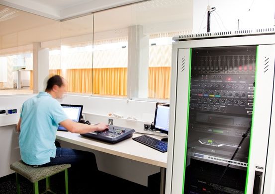 Ein Mann sitzt an einem Schreibtisch mit zwei Monitoren und einer Mischkonsole, wahrscheinlich in einem Büro oder Kontrollraum, und trägt ein hellblaues Hemd.