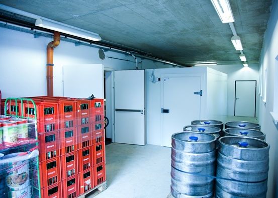 Ein Lager mit weißen Wänden und Decke, das rote Kisten mit Flaschen, Stahlfässer und weiße Türen speichert, beleuchtet von Deckenleuchten.