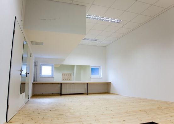 Ein leerer Raum mit weißen Wänden und Holzboden. Ein langer Schreibtisch mit einem Heizkörper und zwei Spiegeln ist an einer Wand positioniert. Zwei Fenster befinden sich an der gegenüberliegenden Wand. Die Decke hat ein weißes Fliesenmuster und zwei Leuchtstofflampen.