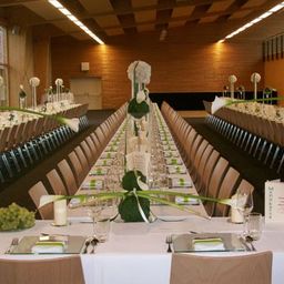 Ein großer Speisesaal ist für eine formelle Veranstaltung vorbereitet, mit weißen Tischdecken, Gläsern und grünen Blumensträußen als Mittelpunkt. Der Raum hat Holzwände und Decken mit Beleuchtung.