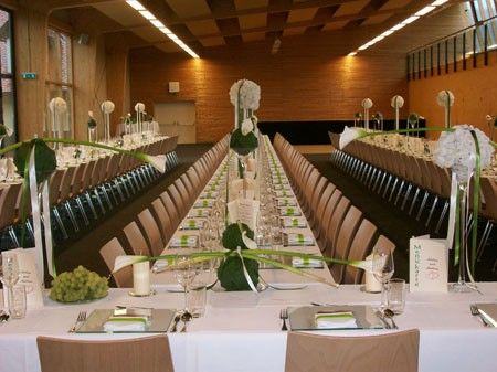 Ein großer Speisesaal ist für eine formelle Veranstaltung vorbereitet, mit weißen Tischdecken, Gläsern und grünen Blumensträußen als Mittelpunkt. Der Raum hat Holzwände und Decken mit Beleuchtung.