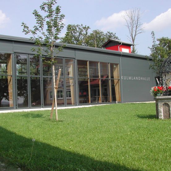 Ein Gebäude mit großen Glasfenstern heißt Kulmlandhalle. Vor ihm steht ein Baum, und rechts befindet sich eine Blumenvase.