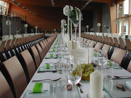 Ein langer Banketttisch mit weißen Tischdecken, Gläsern, Besteck und grünen Blumen. Der Tisch ist für eine Veranstaltung in einem modernen Bankettsaal gedeckt.