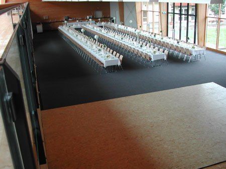 Ein leerer Bankettsaal mit langen Tischen und Stühlen, jeder mit weißen Tischdecken und Blumen besetzt. Der Raum ist geräumig mit Glasfenstern und modernem Design.