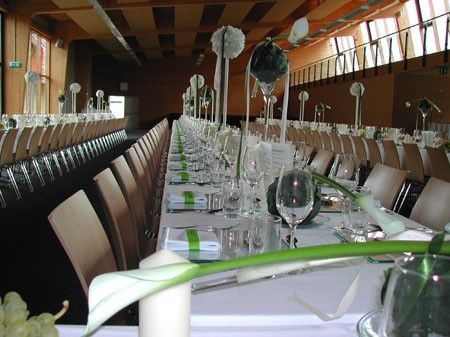 Ein großer Speisesaal mit langen Tischen, die für eine besondere Veranstaltung vorbereitet sind. Die Tische sind mit grünen Bändern, weißen Blumen und hohen weißen Kerzen geschmückt. Jede Tischdecke beinhaltet ein Weinglas und eine gefaltete Serviette.