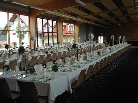 Ein großer Bankettsaal mit langen Tischen, die mit weißen Tüchern bedeckt sind. Jeder Tisch ist mit Tellern, Servietten und Blumen gedeckt. Der Raum hat hohe Decken und große Fenster.