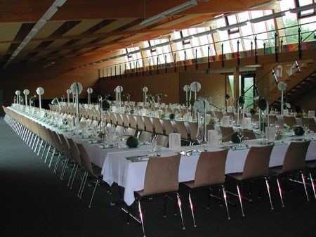 Ein großer Bankettsaal mit Reihen von Tischen und Stühlen, geschmückt mit weißen Tischdecken und dekorativen Tischmitte, unter einer Decke mit großen Fenstern und dekorativen Geländern.