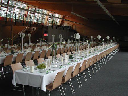 Ein großer Bankettsaal mit langen, in Weiß gedeckten Tischen, jeder mit Blumenschmuck in der Mitte. Stühle sind um die Tische herum angeordnet, und der Raum ist gut beleuchtet mit großen Fenstern und Deckenleuchten.
