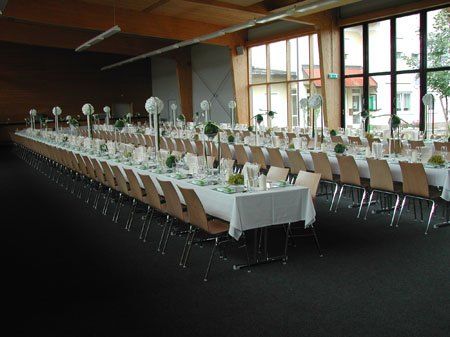 Ein großer Speisesaal mit langen Tischen, die mit weißen Tischdecken bedeckt sind, Metallstühlen und dekorativen Blumenarrangements. Große Fenster lassen natürliches Licht in den Raum.