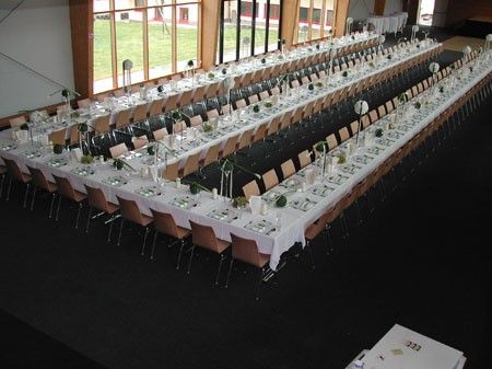 Ein großer Speisesaal ist für eine formelle Veranstaltung vorbereitet, mit langen Tischen, die mit weißen Tüchern bedeckt sind und mit grünen Pflanzen-Zentrenstücken geschmückt sind. Der Saal hat ein modernes Design mit Glaswänden, die Blicke auf ein Grasfeld bieten.