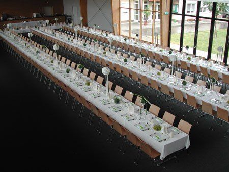 Eine Vogelperspektive eines großen Speisesaals mit langen Tischen, die für eine Veranstaltung vorbereitet sind. Jeder Tisch hat weiße Tischdecken, Blumensträuße als Tischdekoration und mehrere Tischplätze. Der Raum hat große Fenster mit Blick auf eine Rasenfläche.