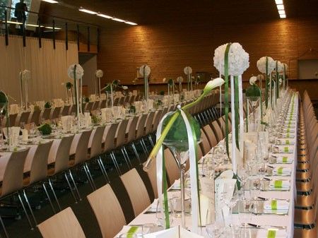 Ein langer Esstisch ist mit weißen Tischdecken, Gläsern und Blumenarrangements gedeckt. Die Stühle sind auf beiden Seiten angeordnet, und der Raum hat Holzwände und Deckenleuchten.