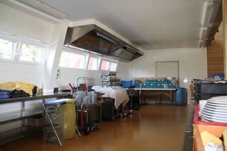 Eine leere Küche mit Holzböden, weißen Wänden und großen Fenstern. Verschiedene Gegenstände sind verstreut, einschließlich Lagerboxen, ein Trolley und ein Fass.