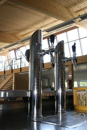 Eine Küchenbereich mit zwei Edelstahl-Bierzapfhähnen und einem gelben Korb. Die Decke ist aus Holz mit großen Fenstern und einem Feuerlöscher.