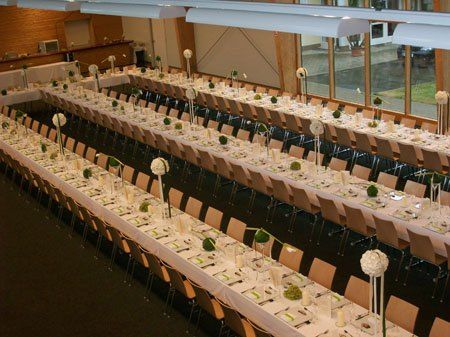 Ein großer Speisesaal ist mit weißen Tischdecken, Blumen und Silbergeschirr dekoriert. Der Raum hat Glaswände, Holzbalken und Deckenbeleuchtung.