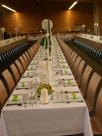 Ein langer Tisch ist für eine Essensveranstaltung gedeckt. Jeder Platz ist mit grünen Bändern und weißen Blumen geschmückt. Der Tisch ist mit einer weißen Tischdecke bedeckt.