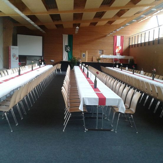 Ein Bankettsaal mit langen Tischen und Stühlen, jeweils mit weißer Tischdecke und rotem Tischläufer. An den Wänden hängen Flaggen und eine Leinwand ist montiert. Der Raum ist leer.
