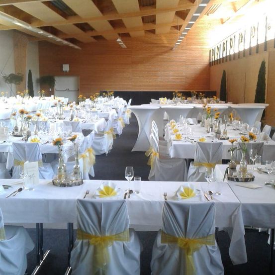 Ein Bankettsaal mit langen weißen Tischen und Stühlen. Jeder Tisch ist mit weißen Tischdecken und gelben Bändern dekoriert. Gelbe Blumen in Vasen stehen auf den Tischen. Der Raum hat Holzwände und eine hohe Decke.
