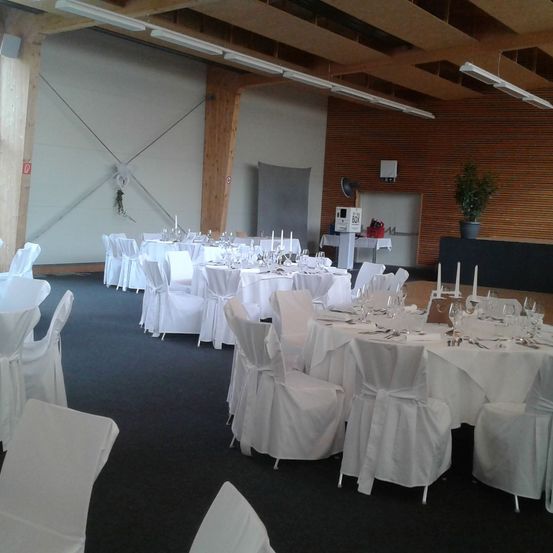 Ein Bankettsaal ist für eine Veranstaltung mit weiß bezogenen Tischen und Stühlen vorbereitet. Holzbalken stützen die Decke, und es gibt Lampen, einen Topfpflanzen und eine Kiste auf einem Ständer.