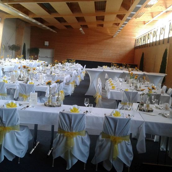 Ein Bankettsaal mit weißen Tischdecken und in Reihen angeordneten Stühlen. Die Tische sind mit Sonnenblumen und gelben Bändern geschmückt. Der Raum hat Holzwände und Decken mit heller Beleuchtung.