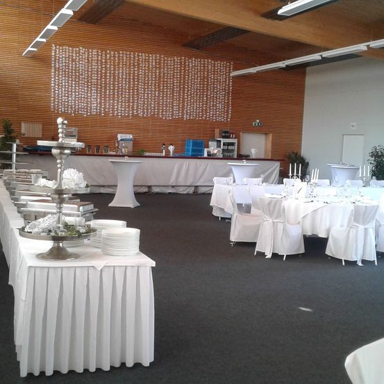 Ein Bankettsaal mit einer Holzwand und langen Tischen, die für eine formelle Veranstaltung gedeckt sind. Die Tische sind mit weißen Tischdecken und Stühlen geschmückt, und ein Buffettisch ist aufgebaut.