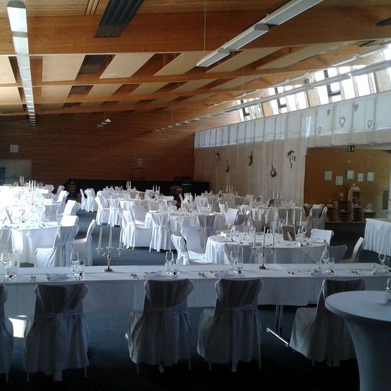 Ein großer Bankettsaal mit weiß gedeckten Tischen und Stühlen, geschmückt mit Kerzen und Weingläsern. Die Decke ist aus Holz mit hängenden Lampen und Fenstern. Der Raum ist geräumig und gut beleuchtet.
