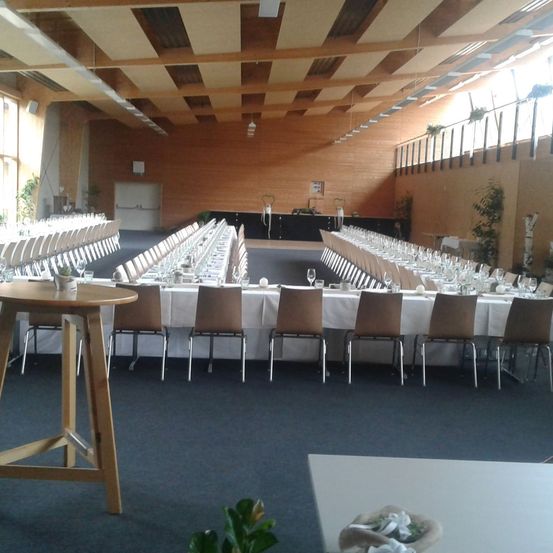 Ein großer Bankettsaal mit Holzwänden und -decke. Reihen von Tischen und Stühlen sind für eine Veranstaltung vorbereitet, mit weißen Tischdecken. Es gibt Topfpflanzen und Beleuchtung.