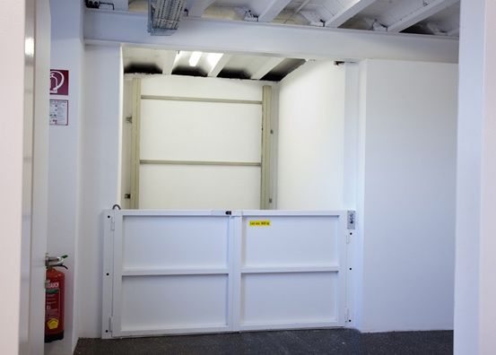 Ein leerer Raum mit weißen Wänden und einer weißen Decke, ausgestattet mit einer weißen Schiebetür und einem roten Feuerlöscher. Es gibt ein kleines Fenster im oberen Teil der Wand.