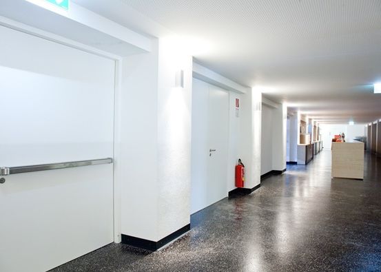 Ein leerer Flur mit weißen Wänden, einem schwarzen Boden und einem roten Feuerlöscher. Es gibt Türen und eine Holztheke.